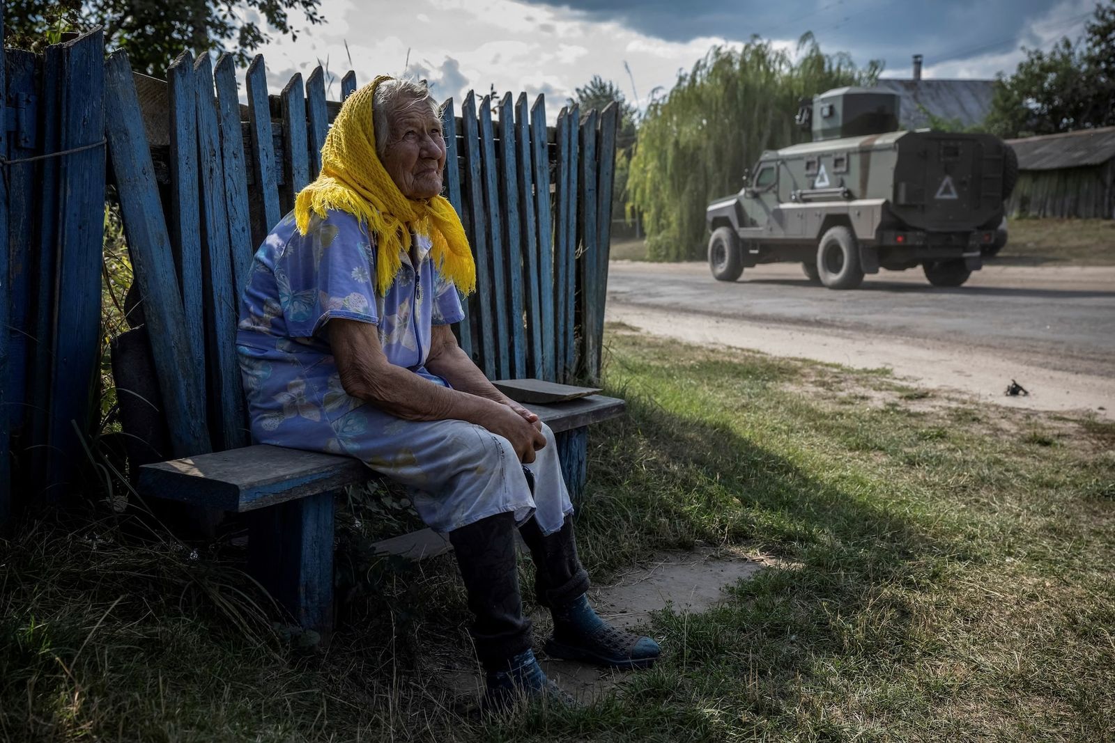 88-летняя жительница Мария возле своего дома, на фоне — военный автомобиль ВСУ, недалеко от российской границы в Сумской области.